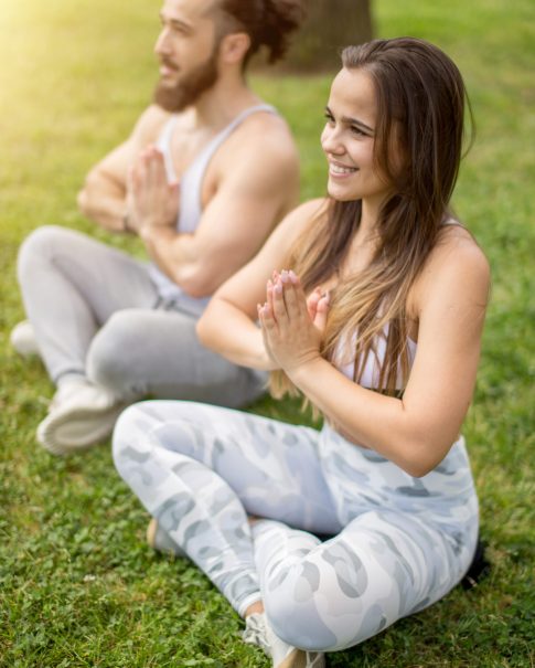 Young couple doing yoga outdoors summer holidays, vacation activities, love and sport lifestyle concept - happy young couple in activewear sitting on green meadow in Lotus position, meditating, feeling comfortoble and free