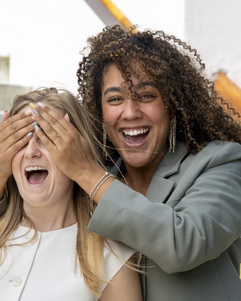 Woman covers friend's eyes with her hands as they both laugh on outdoor steps.